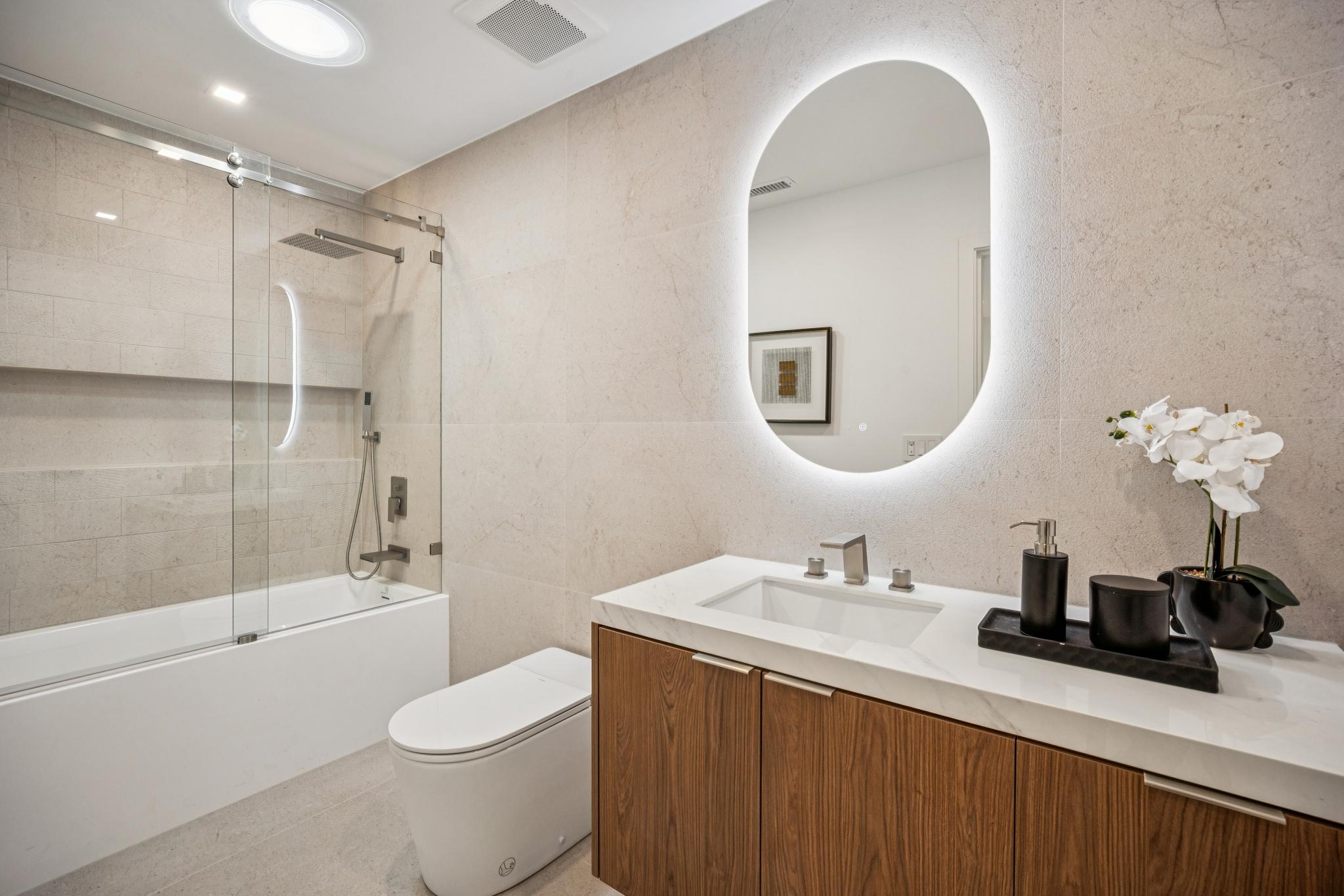 Beautiful luxury bathroom in a Willow Glen custom home, showcasing a floating wood vanity, a glowing LED oval mirror, and a modern glass-enclosed shower and tub combination by Ritz Builders.